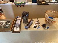 Overview of Native American art and collectibles displayed on a table against a beige wall, including framed art, a clay pot, feather pieces, a wooden artifact, a decorative woven basket, and a small sculpted item.
