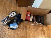 View of box containing various DVDs including Star Trek, Titanic, The Artist, and others. Also visible are remote controls, 3D glasses, and loose discs on the floor.