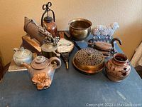 Photo showing assortment including cast iron sheep doorstop, copper bed warmer, stoneware teapots, iron, and other metalware on table.