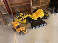 CAT ride-on digger toy and two smaller Tonka truck toys on basement floor with wooden bookshelf in background