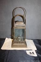 Full frontal view of the vintage kerosene oil lantern placed on a white cloth with a label numbered 34 visible, showing beveled glass panel and metal top.