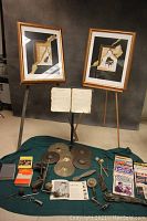 Two framed prints on easels, sheet music held on a stand, cymbals, and assorted music books plus drum accessories laid out on green cloth.
