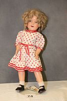 Full body shot showing the doll standing in a cream and red polka dot dress, wearing black shoes and white socks. Some visible wear and damage on limbs.