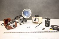 Four vintage cameras displayed on a table with ruler for scale: Kodak Signet 30 with leather case, Kodak Dunfex III with flash, Polaroid Swinger 20, and Vivitar DVR 410 digital camera with accessories.