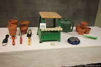 Entire garden lot shown on white tablecloth: clay pots, watering can and wand, sprinkler, gardening tools, ceramic magnet, and garden stool
