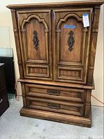 Full front view of the armoire chest showing wood finish, doors, drawers, and metal hardware.