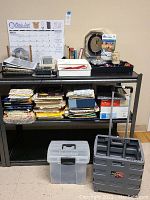 View of black shelf unit holding various office supplies including stacked file folders, boxes, clear tote, and wheeled cart underneath.