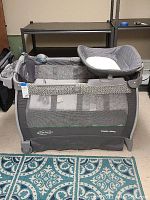 Full front view of gray Graco Pack N Play showing bassinet and attached diaper changer over a blue patterned rug.