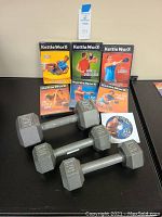 Six Kettle WorX workout DVDs standing upright behind three hexagonal dumbbells (one 20 lb, two 10 lb) on black surface.