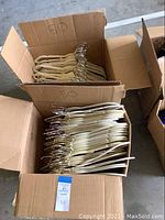 Two cardboard boxes filled with beige and cream felt covered JM Huggable hangers with gold hooks, stacked and organized.