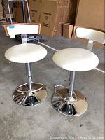 Two contemporary adjustable stools with round off white vinyl seats and backs that have wooden framing, standing on chrome bases with footrests.