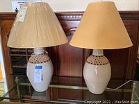 Pair of pottery style lamps with two different parchment look shades sitting side by side on a glass shelf in front of wood paneling.