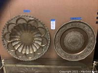 Two round brass pieces on a shelf: larger wall art with scalloped flower pattern and filigree edges, smaller round platter with center floral motif and filigree edge