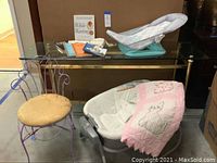 Overview of the lot showing the Graco vibrating baby bed covered with pink and white quilted bear applique blanket, glass table with mommy books and baby bath cradle, and boudoir chair with metal frame and yellow cushion.