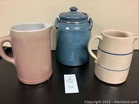 Three pottery items: pink pitcher with handle on left, blue ceramic crock with lid in center, smaller beige pitcher with blue stripe on right
