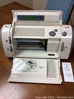 Full frontal view of the white circuit cutting machine with keyboard, display screen, front hatch open showing inside mechanism and manual pages on table.