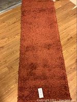 Full top view of burnt orange short shag runner rug laid on wooden floor, showing shape and color, with original label tag visible at one end.