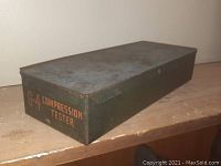 Closed rectangular solid metal box in olive green with orange text 'G-4 Compression Tester'. Surfaces show wear and rust.