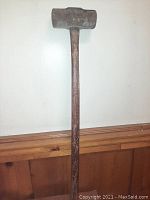 Full view of the large 10lb sledgehammer with wooden handle leaning against a white wall, showing the metal hammer head and wooden handle.