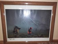 Framed photograph behind glass showing two hunters in orange clothing carrying rifles and elk antlers in a misty wilderness.