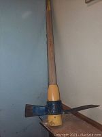 Solid axe and pick tool with a scoop attached, photographed against a wall on a raised surface.