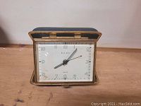 Front view of the older German ELO-Art travel clock showing white dial, black clock hands, and brass case with foldable leather cover.