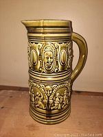 Photo of large glazed pottery pitcher jug showing embossed human faces and figures in olive green glaze.