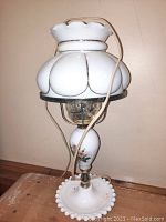 Full side view of a vintage milk glass lamp about 16 inches tall with white scalloped glass lamp shade. Lamp base has hobnail pattern and hand painted floral design with gold trim on the middle section. Electric cord and lamp socket visible.