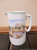Front view of white ceramic pitcher with painted country scene and green rim