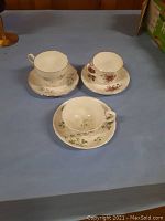 Three bone china tea cups and saucers with different floral patterns and gold accents arranged on a blue surface.
