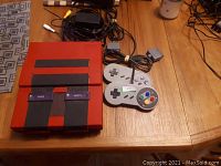 Painted red SNES console on wooden table with two controllers, AC adapter and composite AV cables.