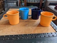Four Fiesta Ware mugs on a table, showing two in peach/apricot, one cobalt blue, and one light blue.