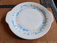 White ceramic cake serving plate with blue and gray mid-century modern utensil pattern around the rim and handles on two sides.