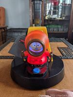 Front view of the lamp showing the detailed Kenny figure in orange parka with magnifying glass over face and light source inside the lamp shade.