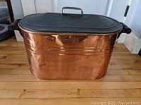 Front view of the antique copper boiler with black tin lid and metal handle, showing its rectangular shape and dented copper surface.