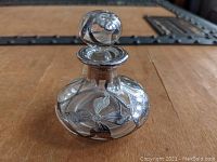 Front angle view showing floral silver overlay decoration on the rounded clear glass body and spherical stopper.