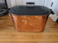 Full view of the antique copper boiler from the front, showing dented hammered copper body and black painted tin lid with wooden handle.