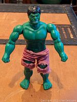 Front view of the green Hulk action figure standing on a brown surface, showing muscular torso, green skin, angry face, and torn purple shorts.
