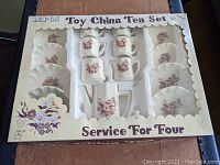 Top view of the full 16-piece vintage Summco porcelain child's tea set in original box with yellowed cellophane cover, showing cups, saucers, plates, teapot, sugar bowl, and creamer arranged in white plastic insert.