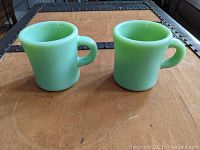 Front view showing two opaque green jadeite glass coffee mugs with large handles, placed side by side on a wooden surface.