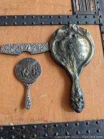 Photo showing all three pieces: large hand mirror, small round hand mirror, and hair comb, arranged on a wooden surface with metal straps.
