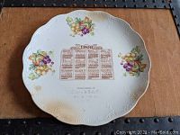 Full view of the 1908 calendar porcelain plate with fruit decorations on a wooden surface.