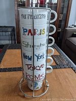 Front view of the six stacked coffee mugs depicting the cities San Francisco, Paris, Shanghai, New York, Tokyo, and Rome in colorful text, held in a metal rack.