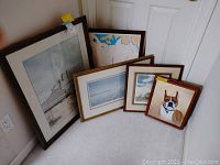 Five framed pictures on floor leaning against wall including ocean and lighthouse scenes, a coastal map, and boxer dog portrait.