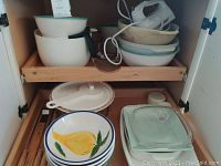 Cabinet shelf with stacks of ceramic mixing bowls, mostly white with green rims, stacked in sets on left and right. Electric hand mixer with cord visible at back center, resting on bowls.