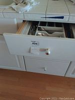 Opened drawer showing various spoons, forks, and butter knives in a white plastic organizer, plus additional utensils like a pizza cutter and sharpening stone on the side.