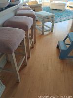 Photo showing four upholstered bar stools with pinkish-purple seats and white painted wooden legs aligned at a kitchen counter, plus one square stool with a blue patterned fabric seat in the background