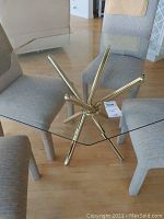 Top view of square clear glass tabletop with brass metal legs and beige upholstered chairs around it. Shows glass surface and metal frame structure underneath.