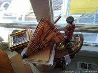 Two framed pictures propped on a table, hand fan style paper umbrella with floral prints, geisha figurine in red attire holding a book, decorative duck figurine, placed on an oval wood table.