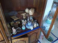 Glass cabinet interior displaying Murphy's by David Winters house figurines, decorative porcelain boxes, framed art, decorative plates, and glass items on two shelves.